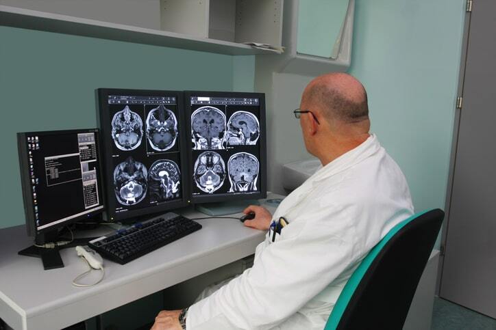 Radiologist analyzing brain MRI scans on multiple monitors using PACS imaging system and RIS software in a medical office setting.