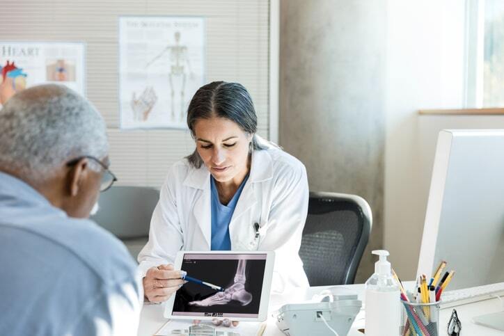 Doctor using a tablet to explain X-ray results to an elderly patient, demonstrating the use of PACS imaging system and RIS software in a medical office setting.