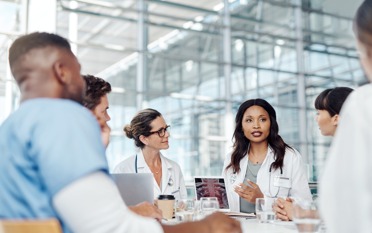 Medical professionals in a discussion around a table in a modern conference room, collaborating and reviewing data on a laptop, illustrating the use of advanced PACS software.